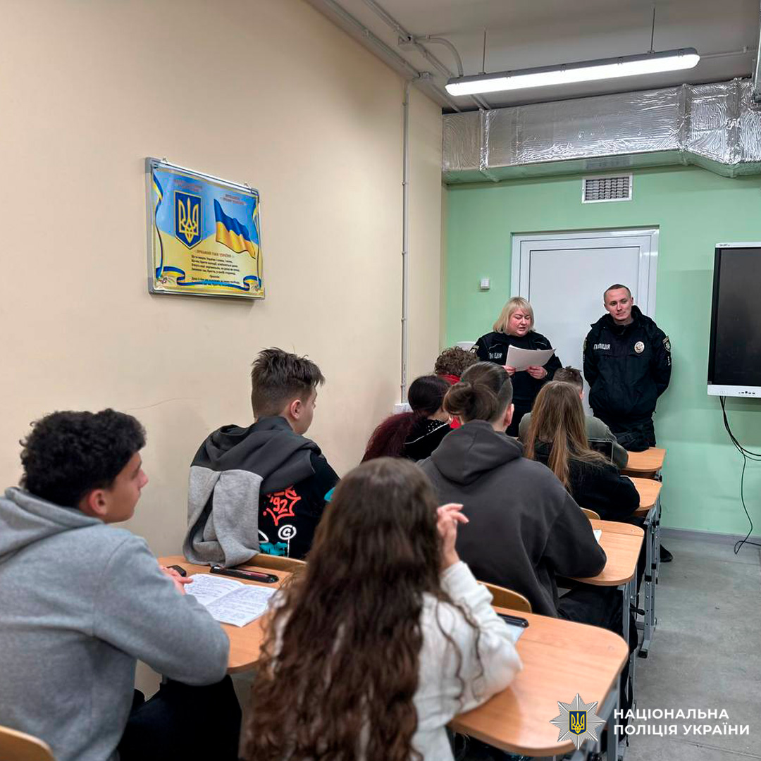Класна кімната, де учні сидять за партами й слухають виступ двох поліцейських у формі.