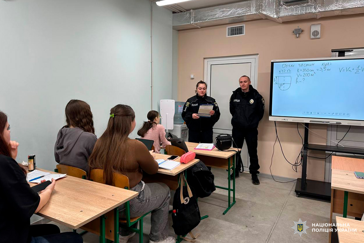 Класна кімната, де учні сидять за партами й слухають виступ двох поліцейських у формі.