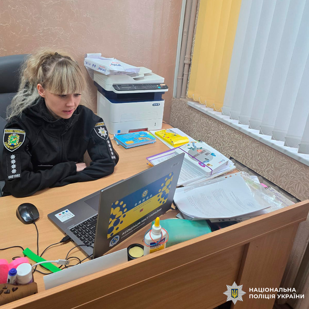 Працівник Національної поліції України у чорній формі з відзнаками, сидить за дерев’яним столом.