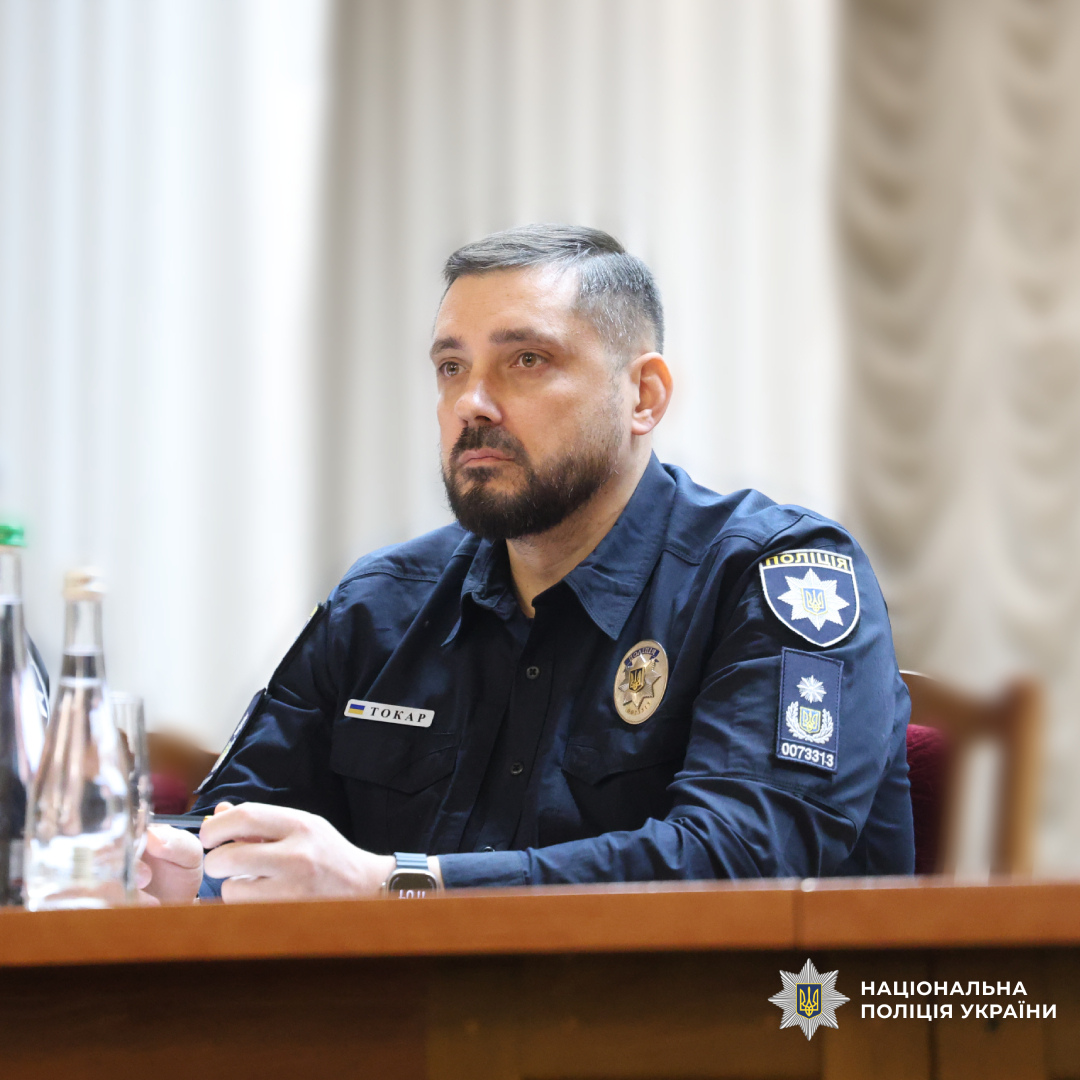 Поліцейський у темно‑синій формі Національної поліції України сидить за столом під час офіційної зустрічі чи конференції.