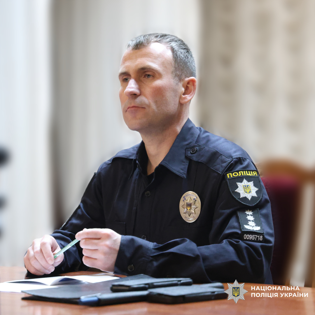 Поліцейський у темно‑синій формі Національної поліції України сидить за столом під час офіційної зустрічі чи конференції.