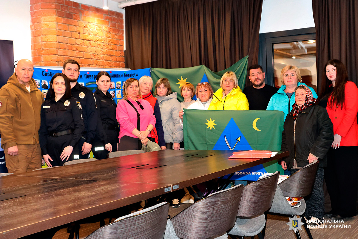 Поліцейські та цивільні громадяни під час урочистого заходу біля банера з фотографіями та символікою Національної поліції України.