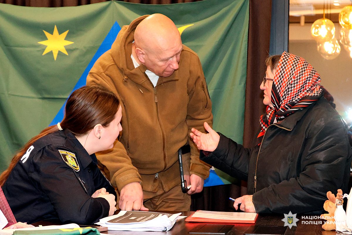 Поліцейський та цивільні учасники за столом під час робочої зустрічі, на тлі прапора з символами.