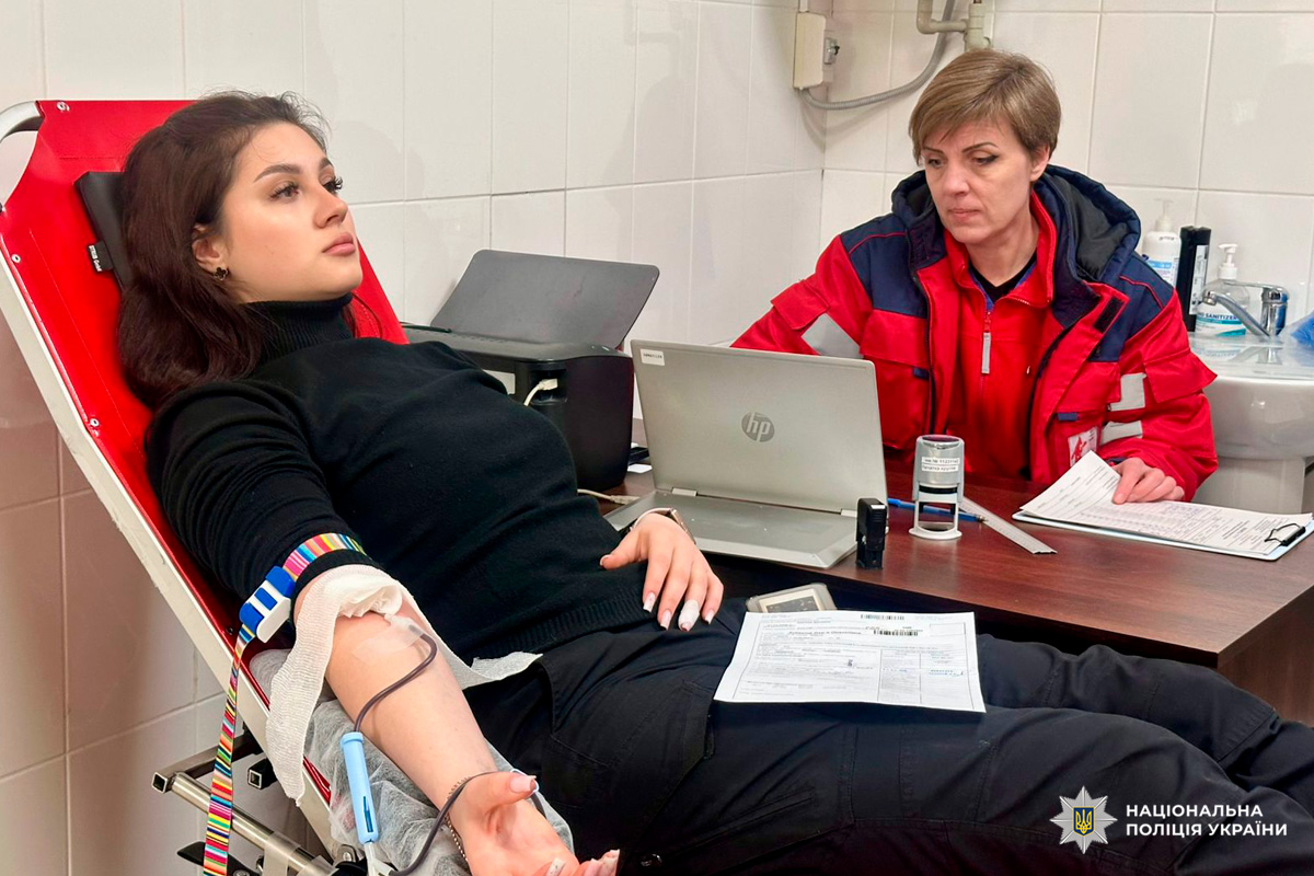 Донор лежить у кріслі з підключеною системою, а медичний працівник у червоній формі веде запис у ноутбуку та працює з документами.