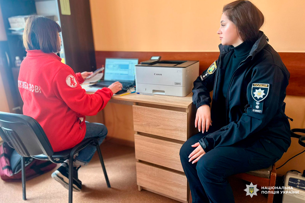 Один учасник у темній поліцейській формі з нашивкою "Поліція" та номером жетона, інший — у червоній куртці з написом "ПТУШКІ Львів" і емблемою із зображенням птаха. На столі — ноутбук, папери та канцелярія.