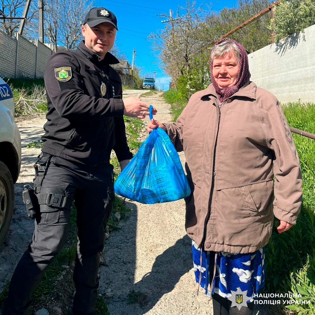 Поліцейський у формі Національної поліції України, з шевронами та спорядженням, стоїть поруч зі службовим автомобілем, частково видимим ліворуч. Жінка у хустці та теплому пальті приймає пакет, що символізує допомогу чи підтримку.