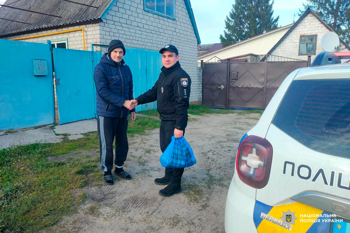 Поліцейський у формі Національної поліції України потискає руку чоловікові, який тримає синій пакет — ймовірно, із допомогою чи передачею. На передньому плані видно службовий автомобіль Renault Duster із написом «ПОЛІЦІЯ» та синьо-жовтими смугами.