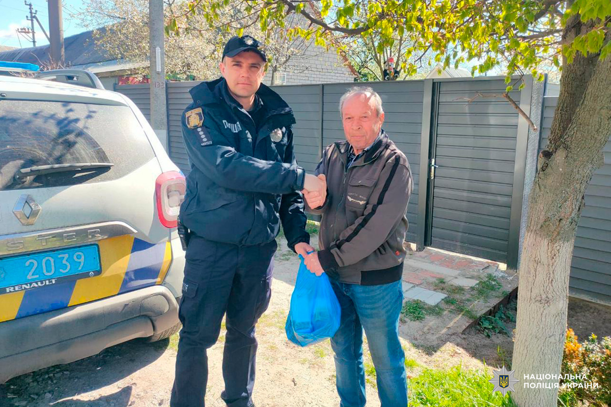 Поліцейський у формі Національної поліції України потискає руку чоловікові, який тримає синій пакет — можливо, із передачею чи допомогою. На задньому плані видно службовий автомобіль Renault Duster із синьо-жовтими смугами, а також весняне дерево з молодим листям.