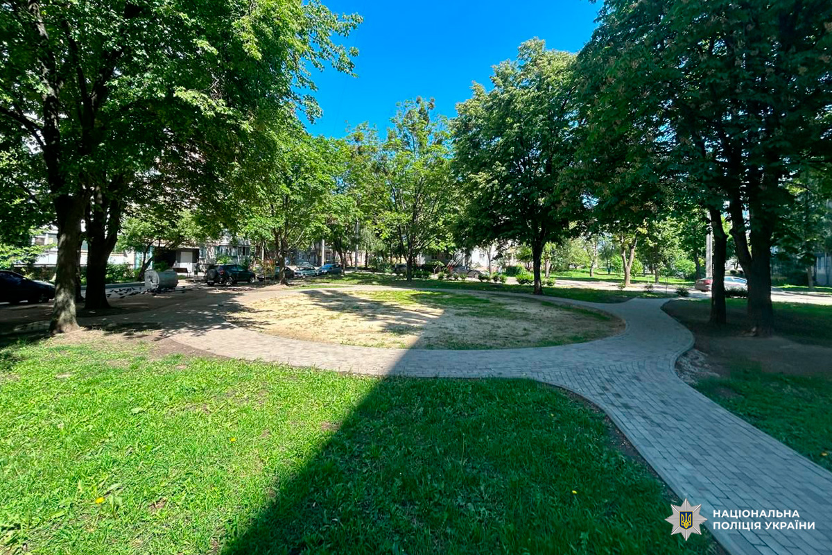 Невеликий сквер або двір із круглою ділянкою трави, оточеною вимощеною доріжкою. Навколо ростуть зелені дерева, що створюють приємну тінь, а на задньому плані видно житлові будинки та припарковані автомобілі. Яскраве синє небо свідчить про сонячний день.