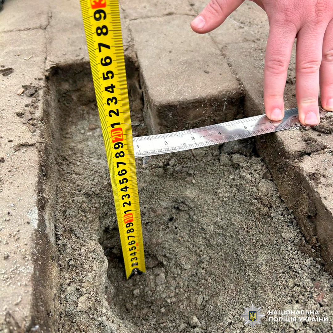 Поліцейський або експерт використовує рулетку та металеву лінійку, щоб зафіксувати параметри заглиблення між бетонними або кам’яними плитками. На зображенні видно руку, що тримає інструмент.