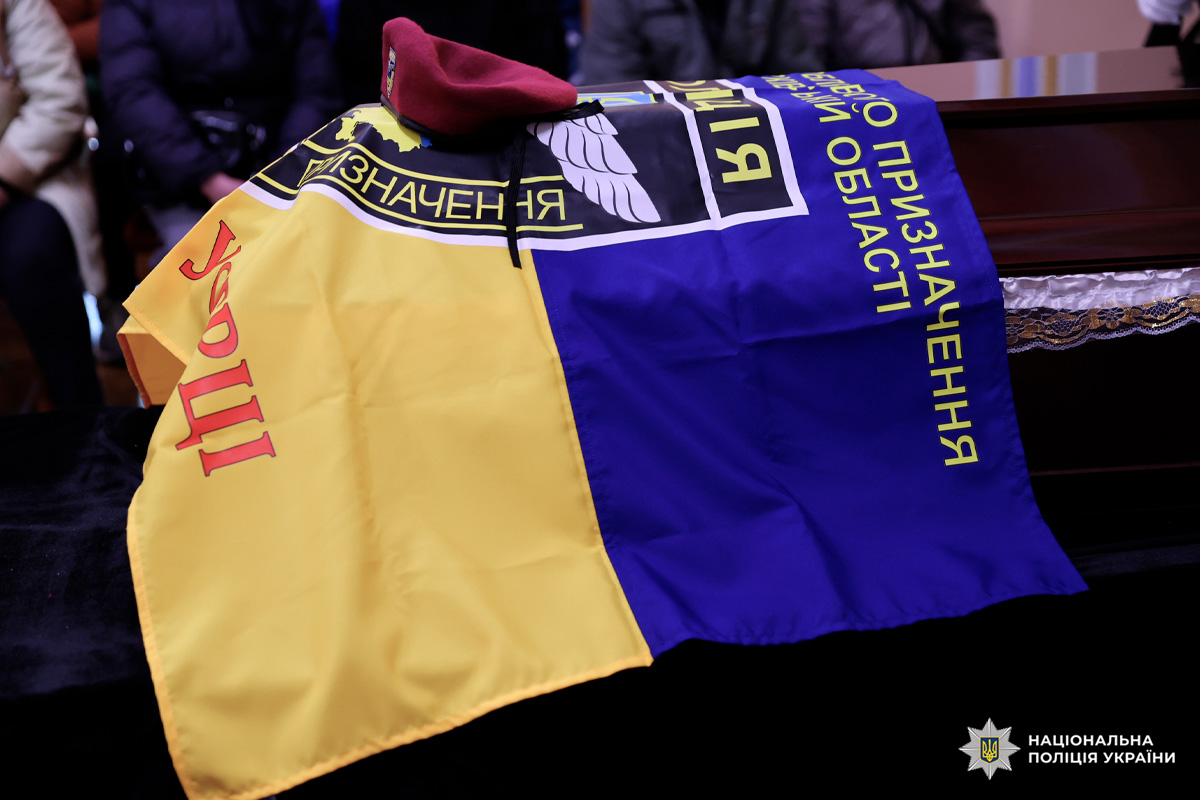 На фото — труна, накрита синьо‑жовтим прапором. На прапорі лежить бордовий берет.