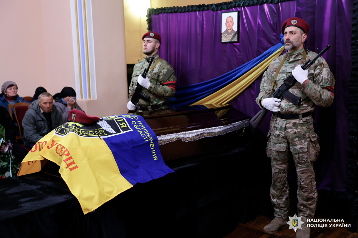 У приміщенні проходить траурна церемонія. У центрі — закрита труна, накрита синьо‑жовтим прапором. По обидва боки стоять двоє поліцейських у камуфляжній формі та бордових беретах, тримаючи зброю у церемоніальній стійці. На задньому плані — драпірування з синьої, жовтої та фіолетової тканини, над труною розміщено портрет загиблого.