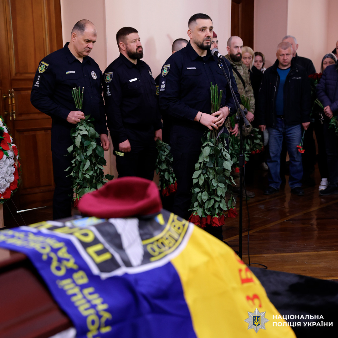 У приміщенні проходить траурна церемонія. На передньому плані — труна, накрита синьо‑жовтим прапором, на прапорі лежить бордовий берет. На задньому плані стоять працівники поліції у формі, тримаючи в руках букети троянд; один із них говорить у мікрофон.