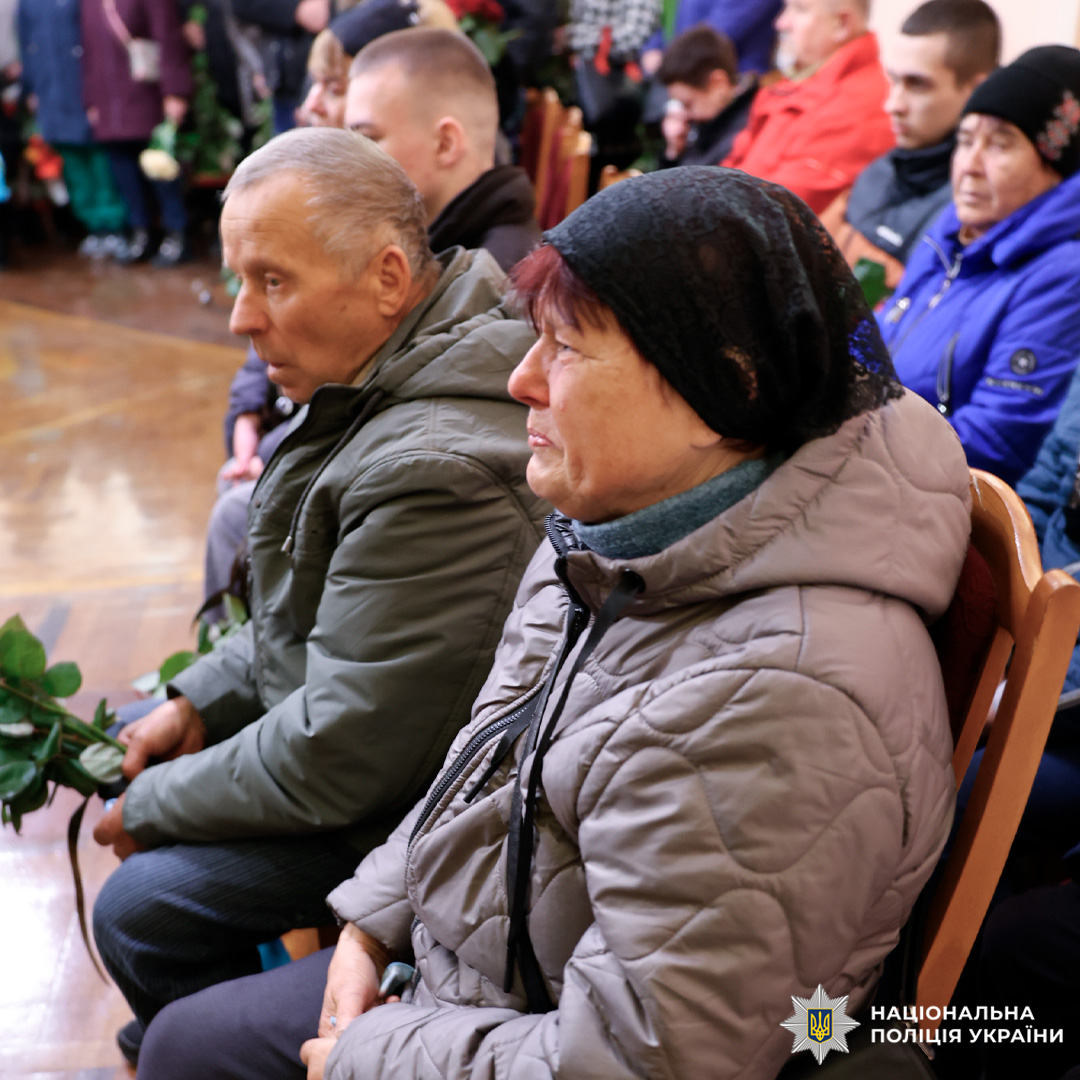 У приміщенні сидять люди, розташовані рядами на дерев’яних стільцях. На передньому плані — літня жінка у сірій куртці та чорній хустці, поруч із нею чоловік у темній куртці. Вони тримають у руках зелені стебла квітів. На задньому плані видно інших присутніх у теплому одязі, що спостерігають за подією.