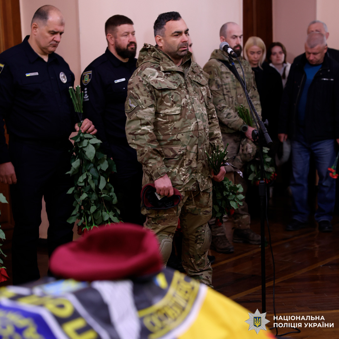 У приміщенні проходить траурна церемонія. На передньому плані — труна, накрита синьо‑жовтим прапором, на прапорі лежить бордовий берет. На задньому плані стоять працівники поліції у формі, тримаючи в руках букети троянд; один із них говорить у мікрофон.