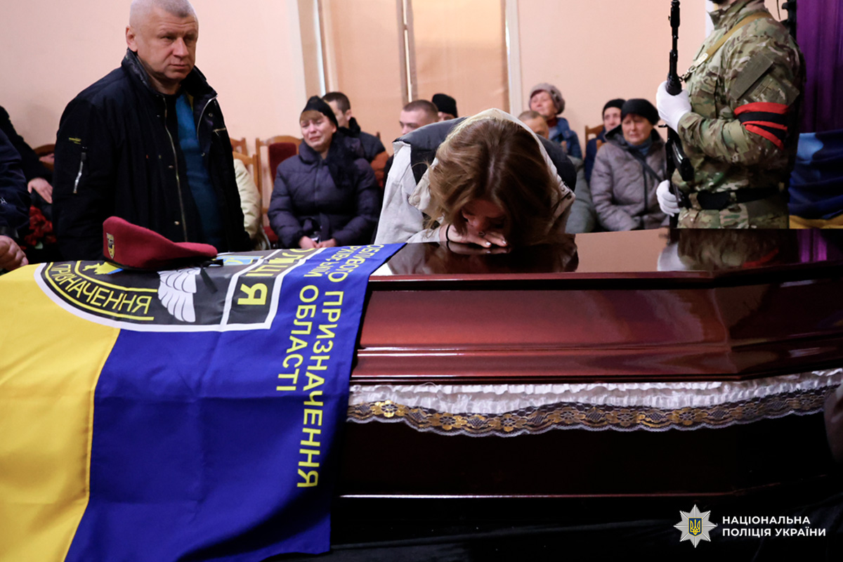 У приміщенні проходить траурна церемонія. У центрі — труна, накрита синьо‑жовтим прапором. На прапорі лежить бордовий берет. Біля труни стоїть чоловік у цивільному одязі, поруч — жінка, схилившись над кришкою труни.