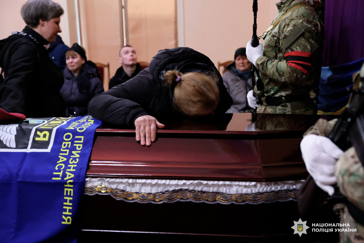 У приміщенні проходить траурна церемонія. У центрі — труна, накрита синьо‑жовтим прапором. На прапорі лежить бордовий берет. Біля труни стоїть чоловік у цивільному одязі, поруч — жінка, схилившись над кришкою труни.