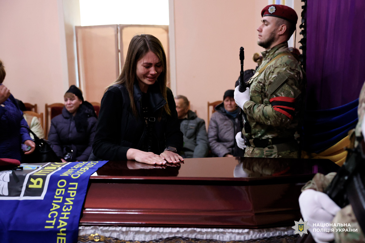 У центрі — жінка, яка схилилася над закритою труною, поклавши руку на її поверхню. Її емоції — глибокий сум і біль втрати. Праворуч стоїть почесна варта у камуфляжній формі з червоними беретами, тримаючи зброю в урочистій позі. На задньому плані — люди, що сидять у скорботі.