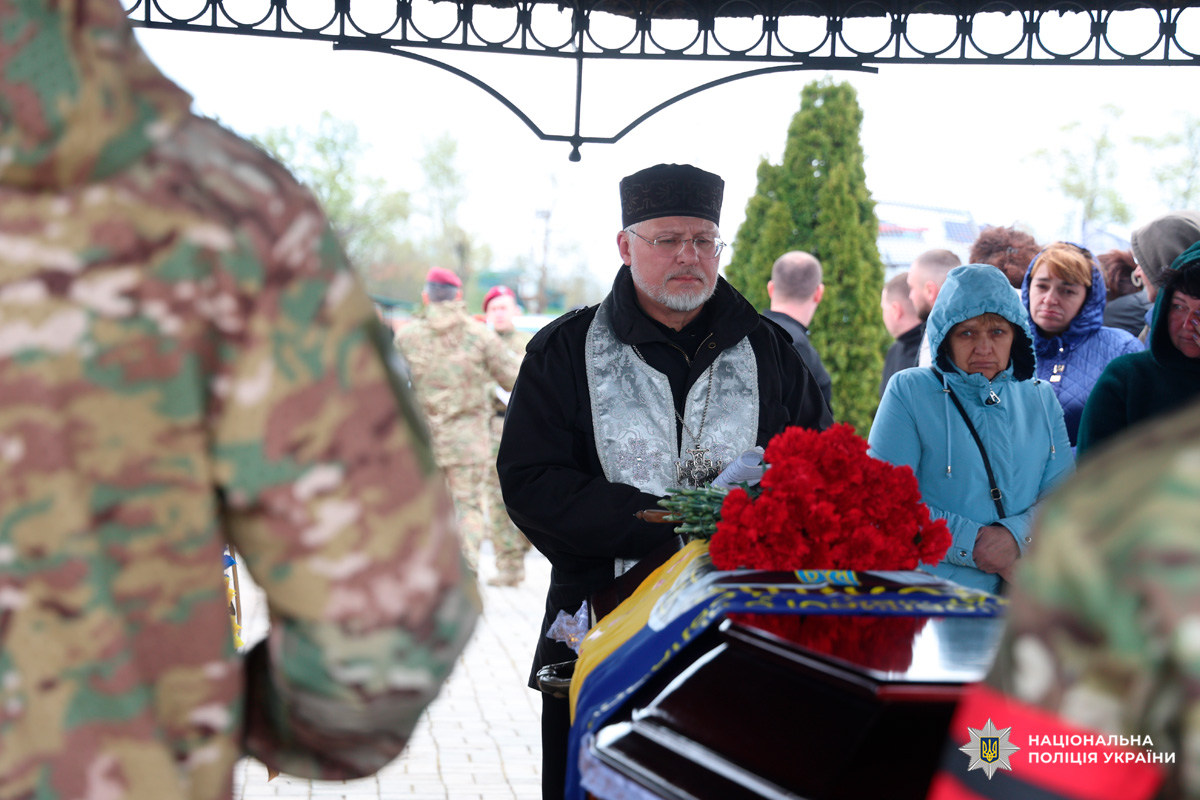 У центрі стоїть священник у чорному головному уборі та сріблястій ризі, тримаючи букет червоних гвоздик біля труни, накритої синьо-жовтим прапором України. Попереду — військовослужбовці у камуфляжі, а позаду — люди в цивільному одязі, які стоять у скорботі.