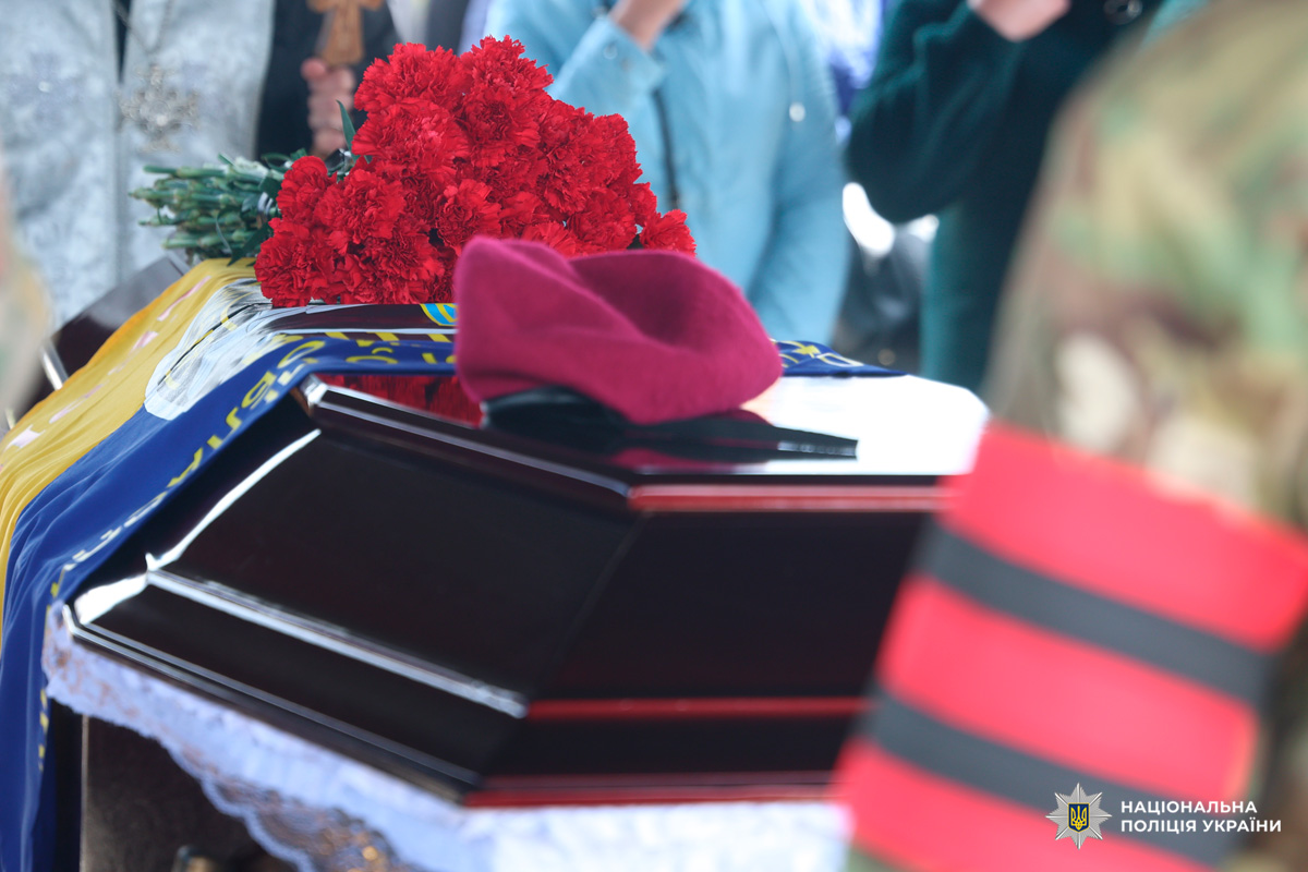 На цьому фото зображено урочистий момент прощання з поліцейським. У центрі — темна лакована труна, накрита синьо-жовтим прапором України. На кришці лежить бордовий берет і букет червоних гвоздик — символи військової честі та пам’яті.