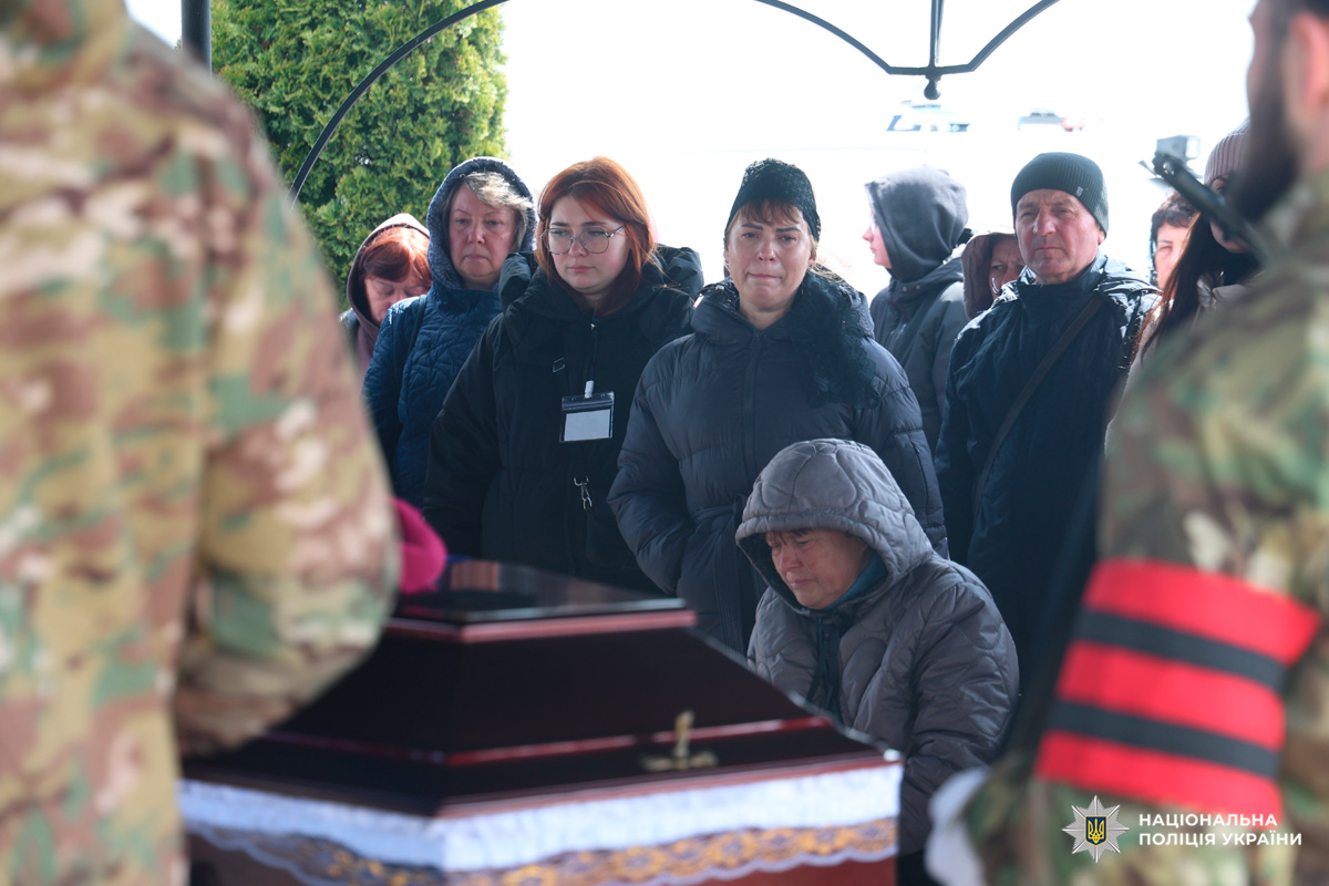 У центрі кадру — темна труна з декоративною окантовкою, перед якою стоять люди у чорних куртках і пальтах, більшість із них із сумними або зосередженими обличчями. Дехто тримає руки схрещеними, одна жінка сидить, схилившись ближче до труни.