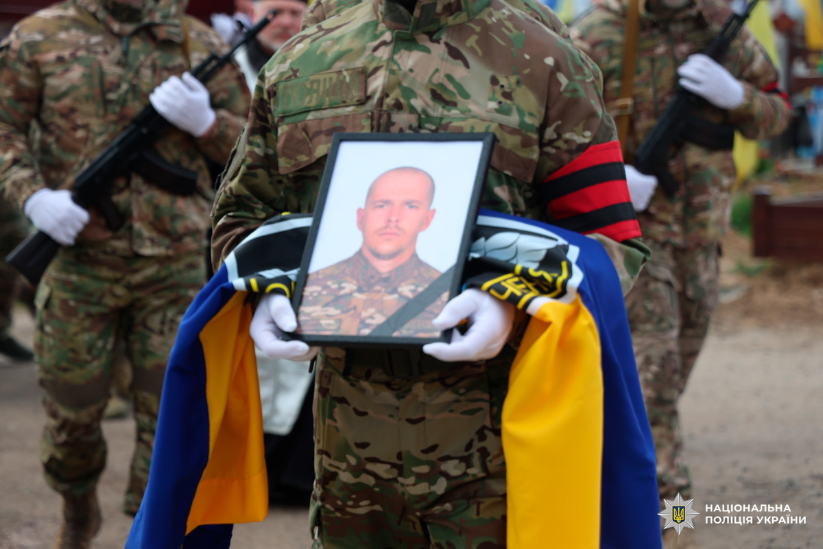 У центрі кадру — військовослужбовець у камуфляжній формі, який тримає портрет загиблого колеги в рамці та складений синьо-жовтий прапор України. На його рукаві — чорна траурна стрічка з червоним елементом, а руки в білих рукавичках підкреслюють офіційність церемонії.