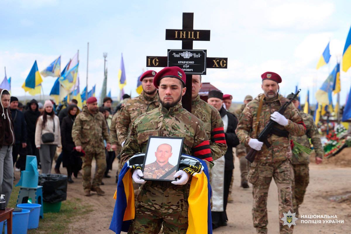 У центрі — військовослужбовець у камуфляжній формі та бордовому береті, який тримає портрет загиблого, складений синьо-жовтий прапор України та стоїть попереду процесії. За ним — інший військовий із дерев’яним хрестом, на якому зазначено ім’я: Кравченко Ігор Ігорович (1996–2024). На задньому плані — колеги у формі, цивільні та численні державні прапори.