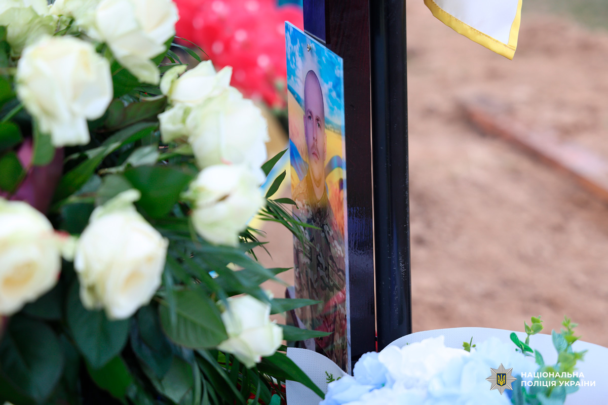 У центрі кадру видно портрет чоловіка у військовій формі на тлі синьо-жовтого прапора, прикріплений до темного хреста. Навколо — квіти: білі троянди, зелене листя та світло-блакитні декоративні елементи, що створюють атмосферу спокою й пошани. На задньому плані — земля, що підкреслює контекст поховання.