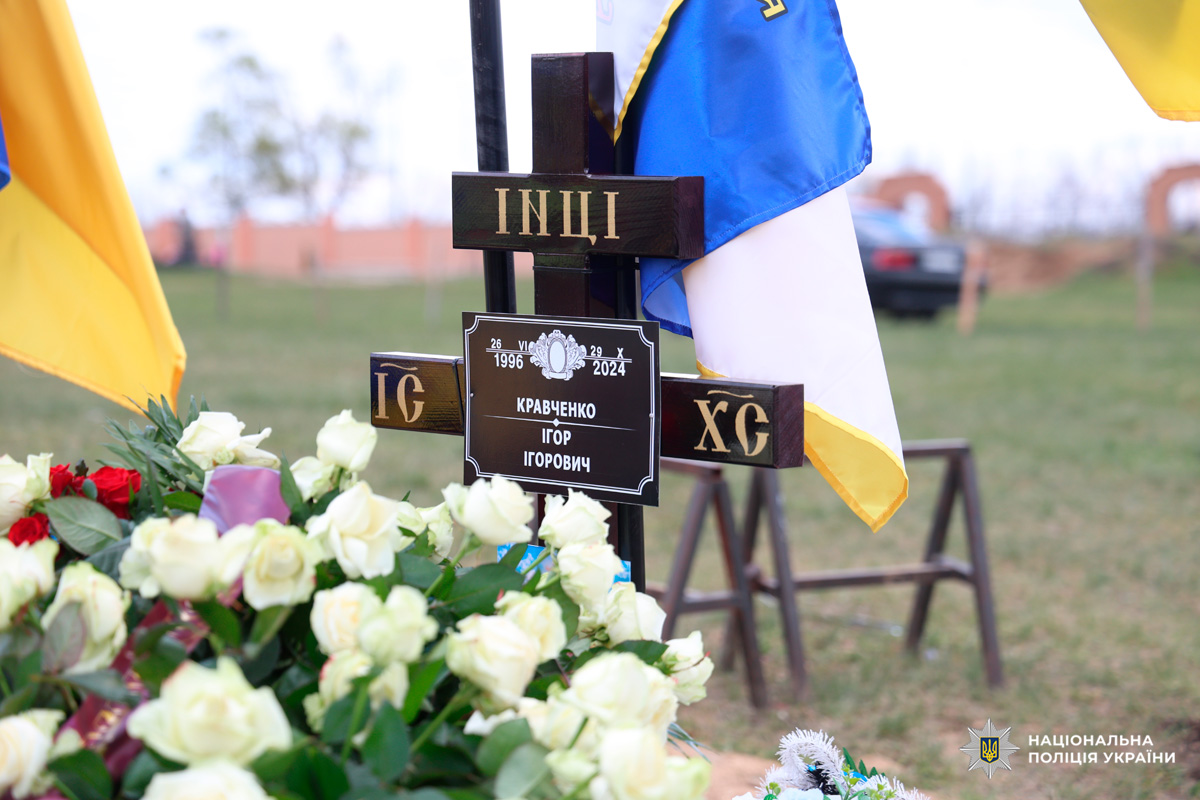 У центрі стоїть дерев’яний православний хрест із золотими літерами, а також табличкою з іменем і датами життя. Навколо — квіти, переважно білі троянди, що символізують чистоту й пам’ять. На задньому плані майорять синьо-жовті державні прапори,