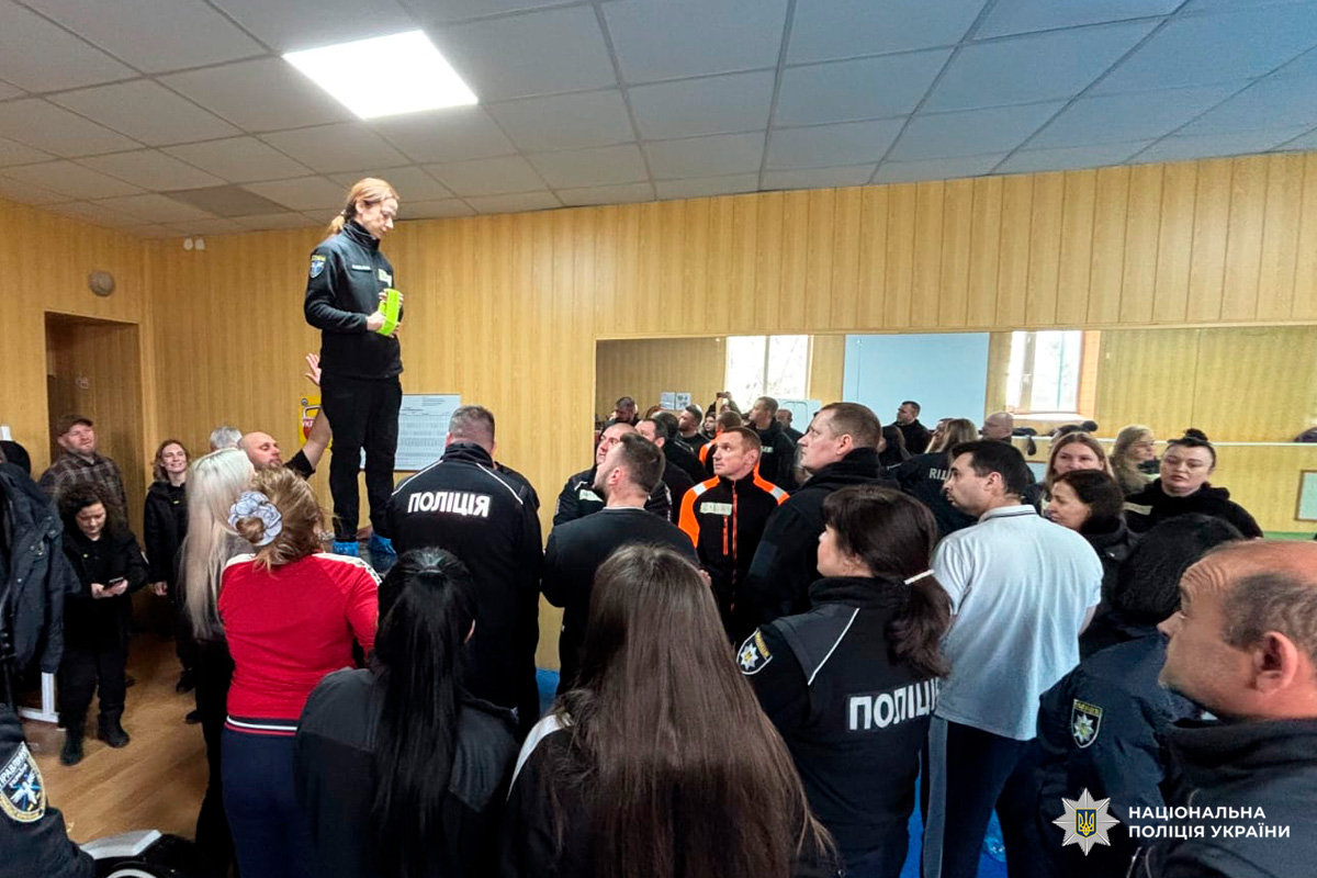 На фото зображено навчальний або командний захід Національної поліції України. У приміщенні з дерев’яними панелями на стінах стоїть група поліцейських у формі з написом «ПОЛІЦІЯ». Один із учасників у спортивному одязі — стоїть на підвищенні, тримаючи зелений предмет у руках, і звертається до присутніх.