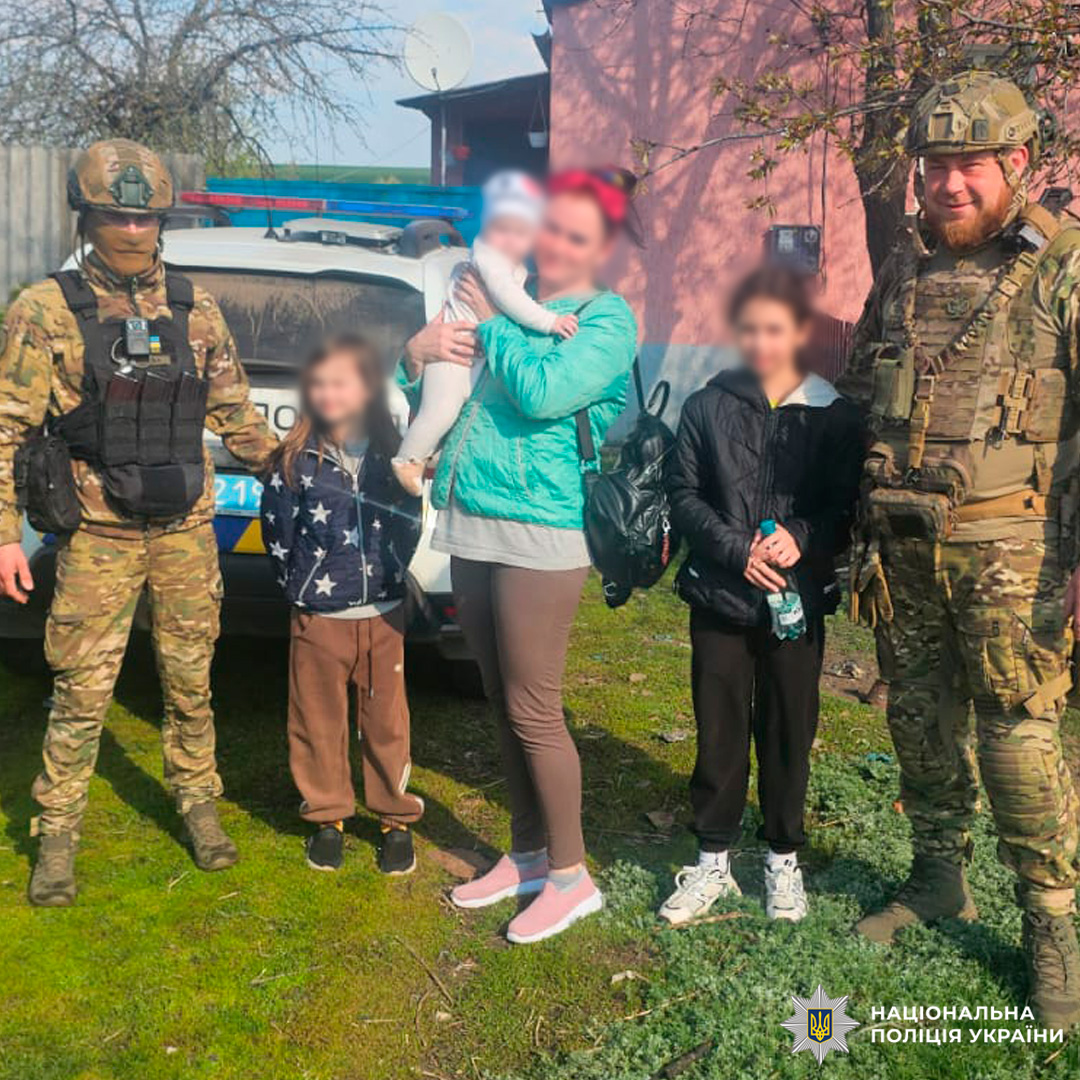 Поліцейські у камуфляжній формі спілкуються з родиною — жінкою з немовлям та двома дітьми — біля службового автомобіля Національної поліції України».