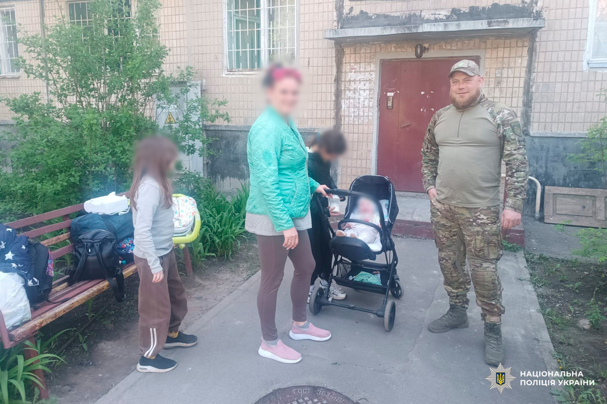Поліцейський у камуфляжній формі спілкується з жінкою та дітьми біля житлового будинку, поруч дитячий візок і лавка з речами.