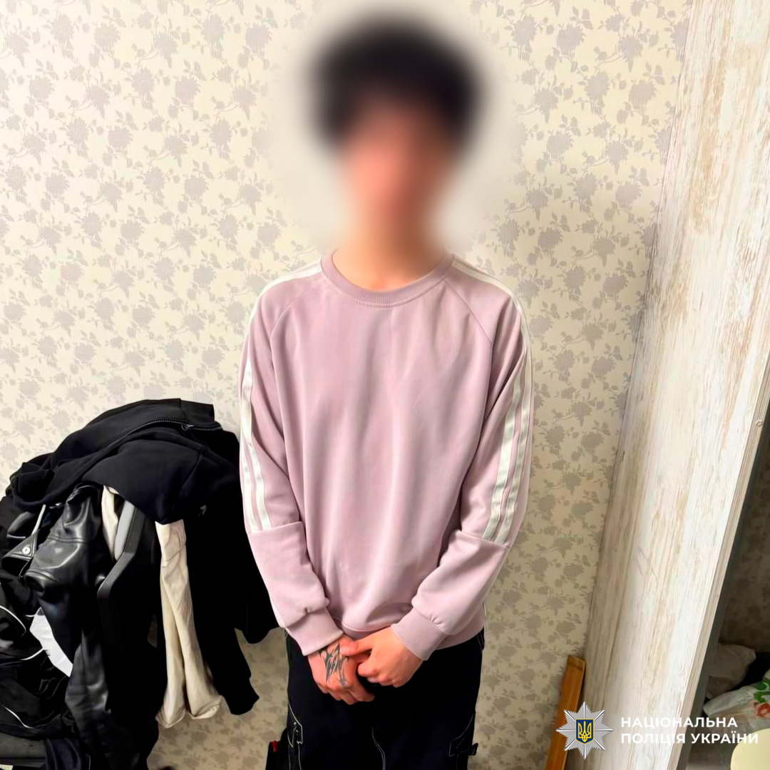 Молодий чоловік у світло‑рожевому светрі стоїть у кімнаті з бежевими шпалерами, поруч із розкиданим одягом і дверним отвором.