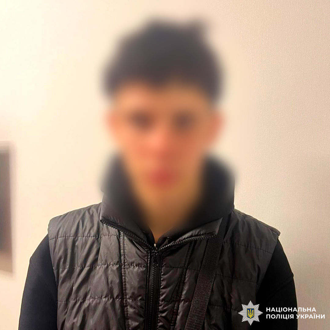 Молодий чоловік у чорній жилетці та худі стоїть біля світлої стіни, дивиться прямо в камеру.