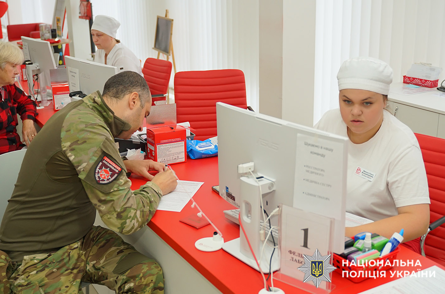 За столом червоного кольору ситиди медсестра перед монітором компёютера. Навпроти неї працівникі поліції у формі заповнює документи.
