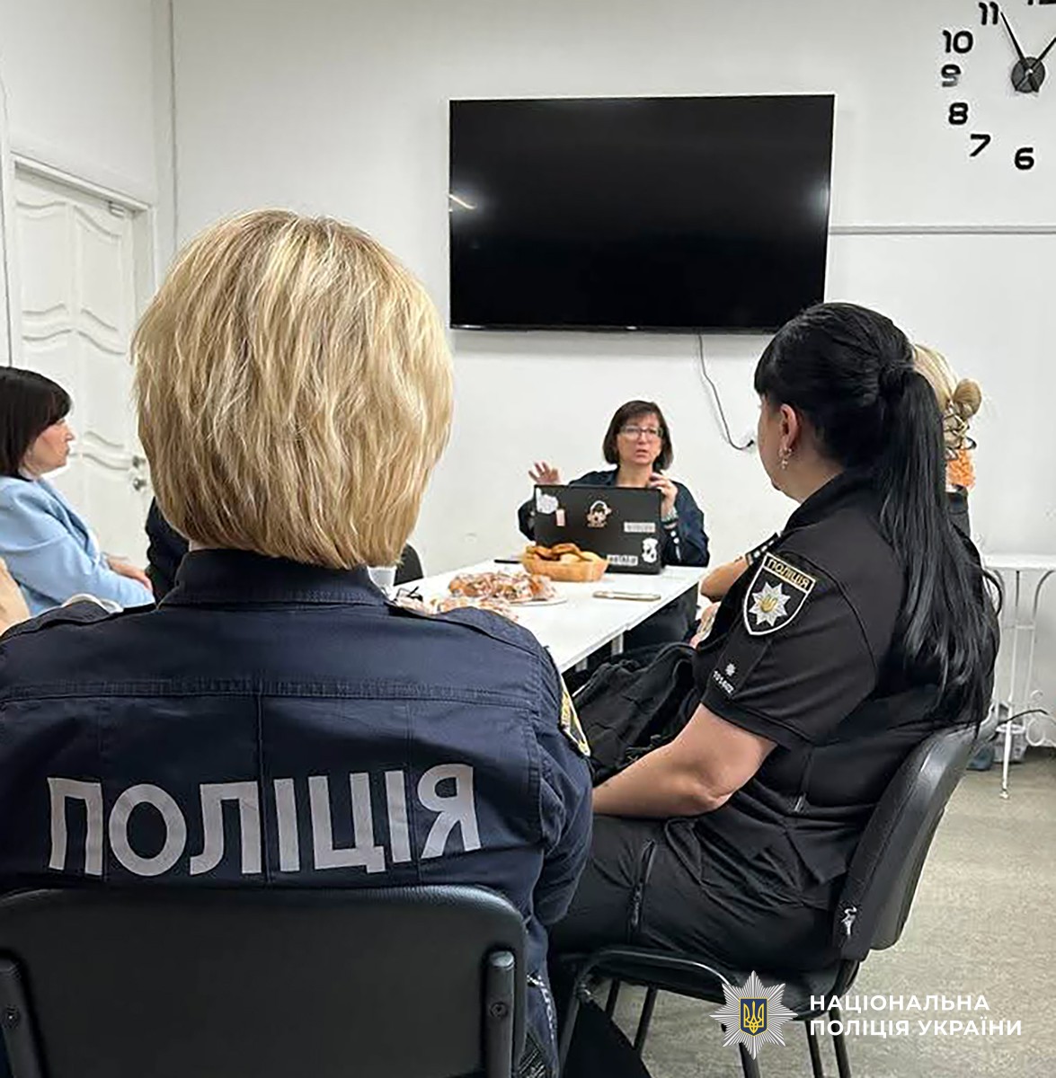 За столом сидять жінки в поліційній формі та цивільному одязі. Одна з них щось розповідає