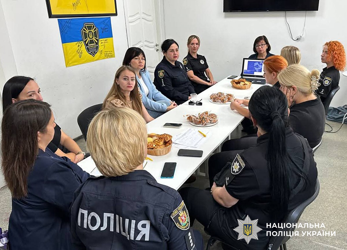 Група жінок у поліційній формі та цивільному одязі сидять за столом, щось обговорюють. На столі - комп'ютер з презентацією та випічка