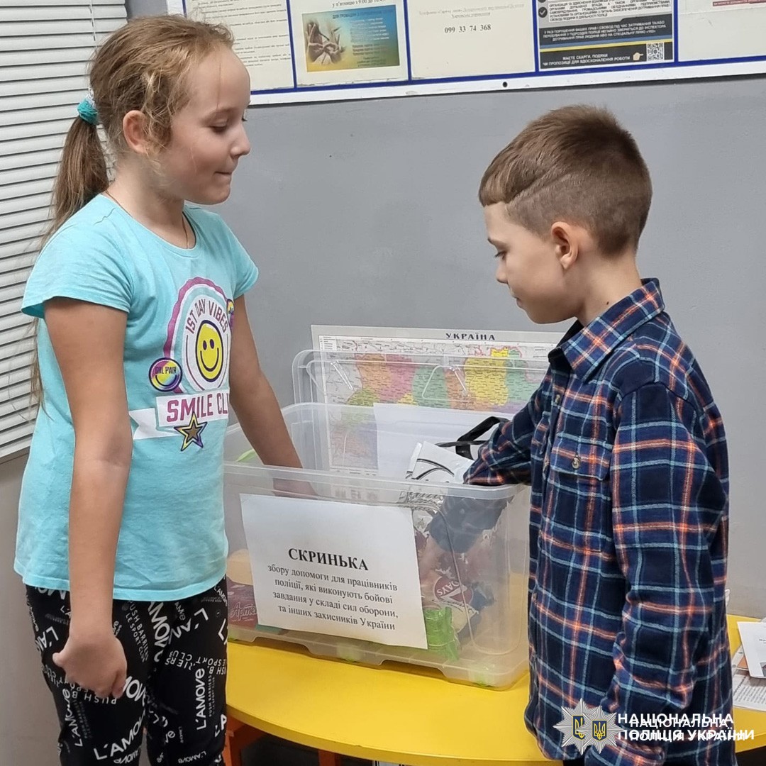 двоє дітей біля коробки з написом "СКРИНЬКА" (коробка для пожертв), поруч мапа України та табличка з інформацією.