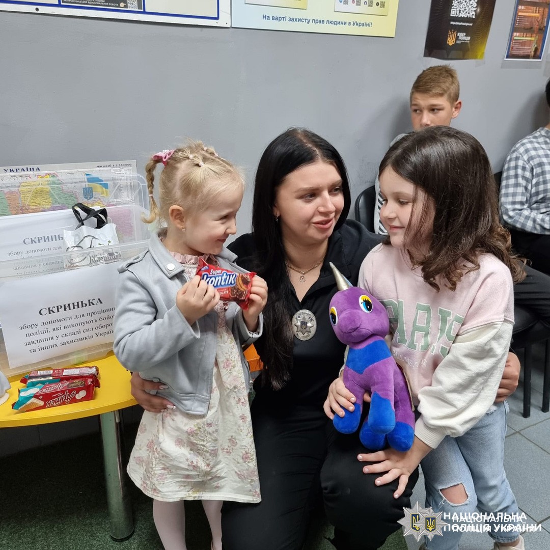 жінка-поліцейська з двома дівчатками тримають плюшевого єдинорога та шоколадку "Snickers" біля стола з цукерками та мапою України.
