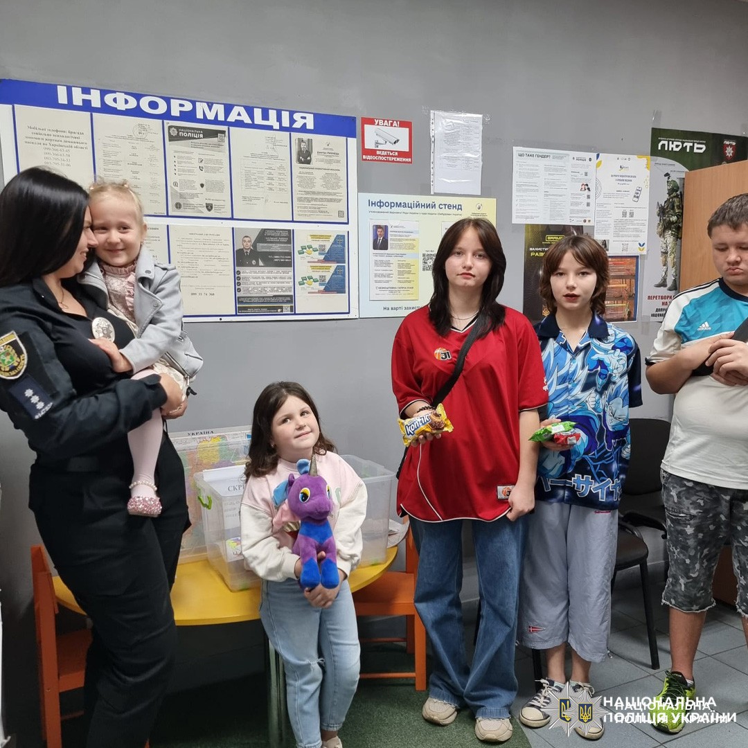 жінка-поліцейська з дівчинкою на руках, троє дітей з плюшевими іграшками та шоколадками біля стола з мапою України та коробкою.