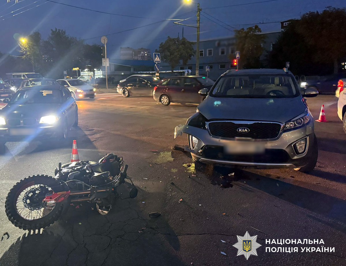 Дорога з інтенсивним трафіком, вечір. на дорозі стоїть пошкоджений автомобіль, а біля нього лежить мотоцикл. Поруч стоять кілька дорожніх конусів