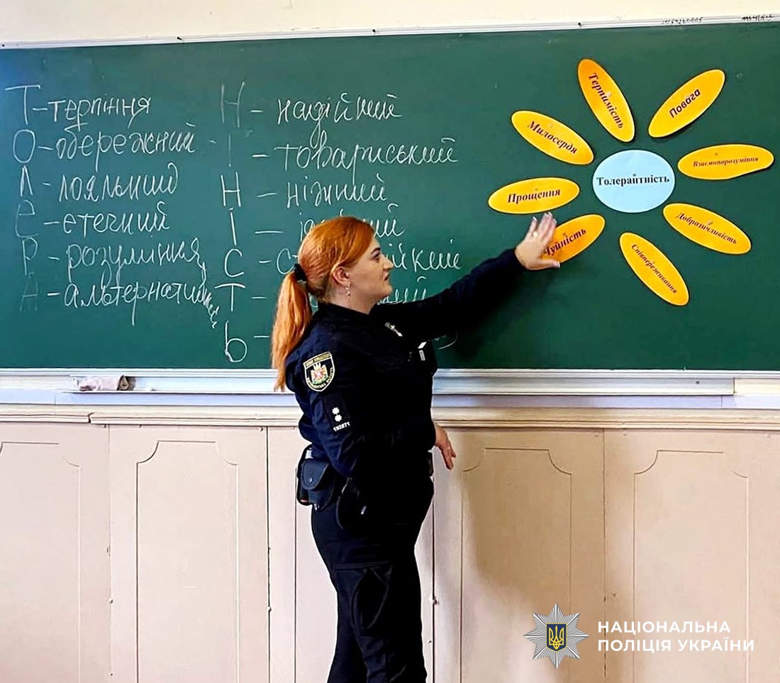 поліцейська стоїть біля дошки перед учнями та вказує ьрукою на пипис, приклеєний на докшу