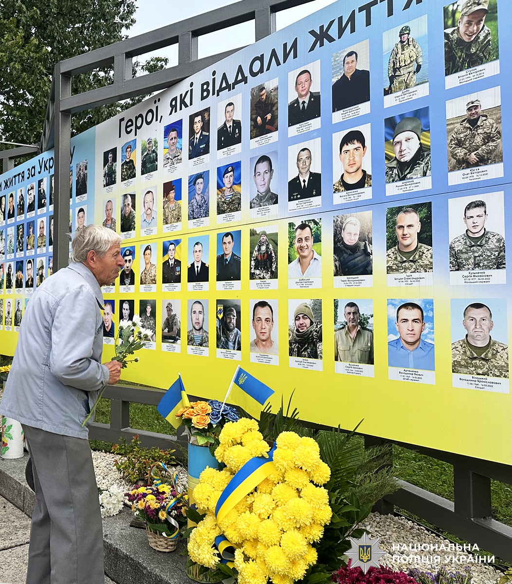 чоловік похилого віку з квітами в руках стоїть перед дошкою зі світлинами полеглих Героїв