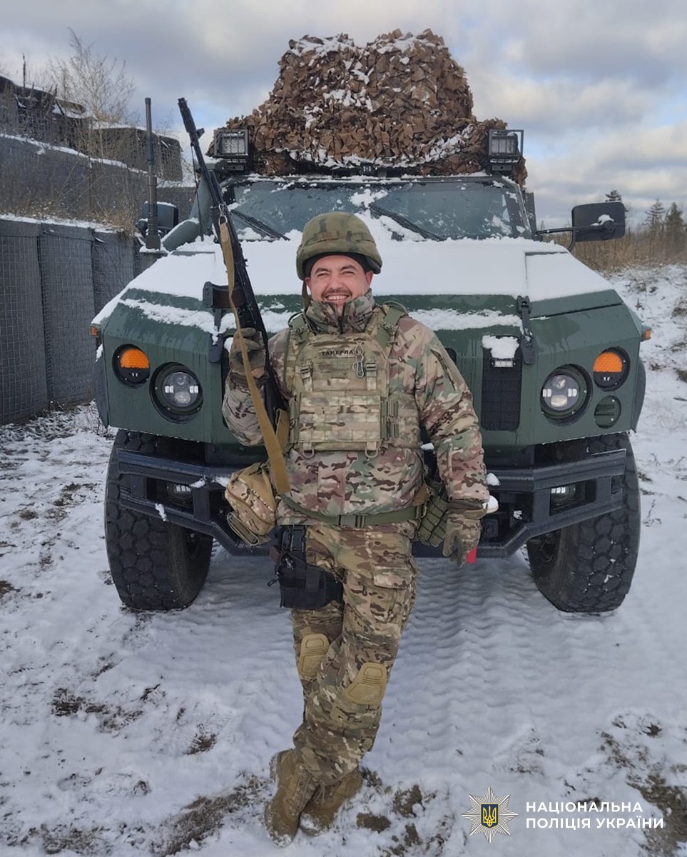 Руслан у повній амуніції з автоматом, на фоні бронеавтомобіля.