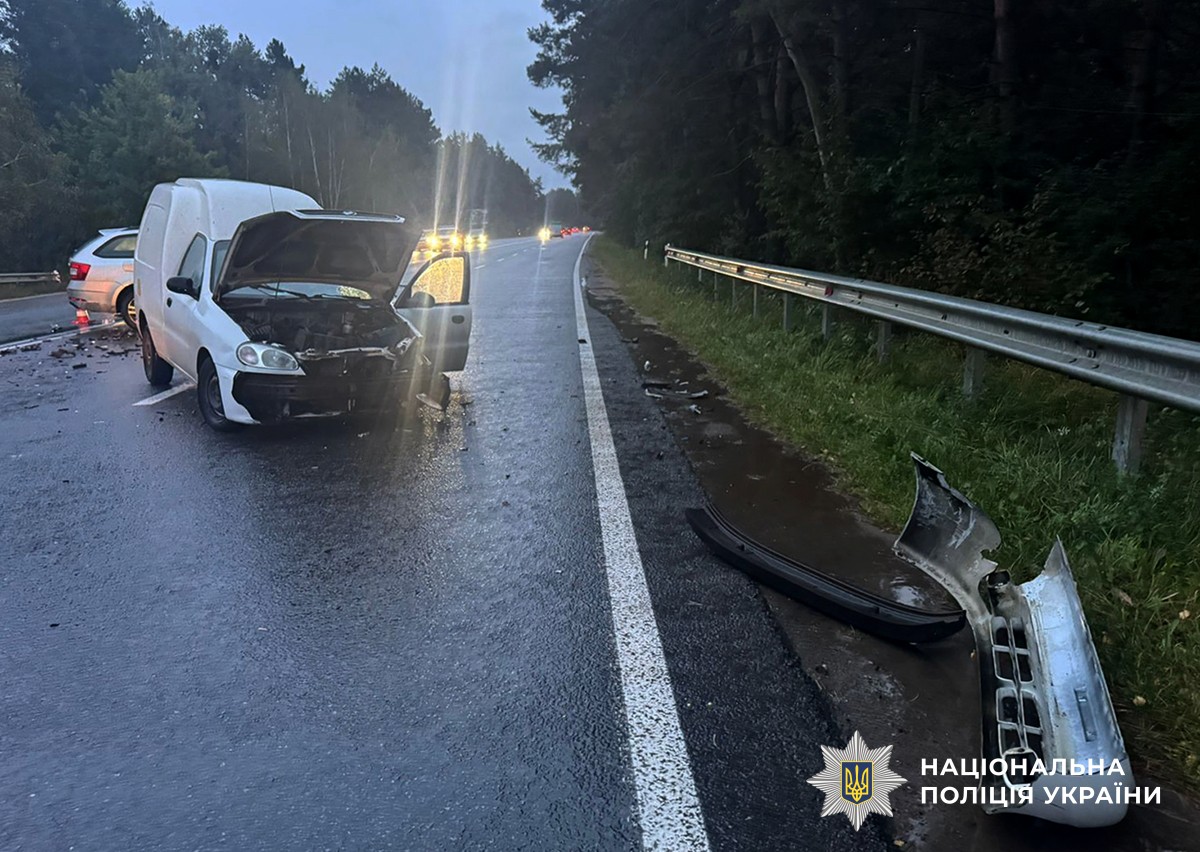 На дорозі стоїть білий автомобіль ЗАЗ із сильно пошкодженою передньою частиною, капот відкинутий догори, дверцята відчинені. На узбіччі лежать частини бампера та уламки авто.