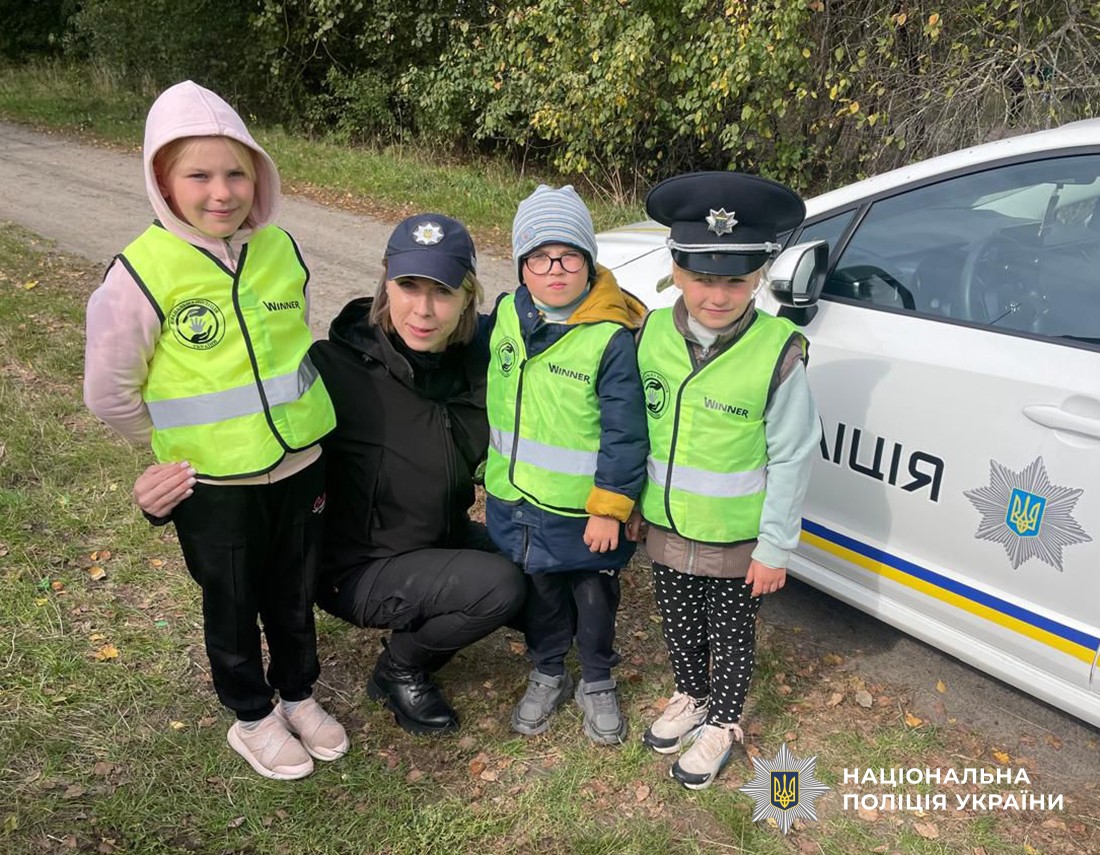 Троє дітей у світловідбивальних жилетах позують біля службового авто, поруч присіла поліцейська.