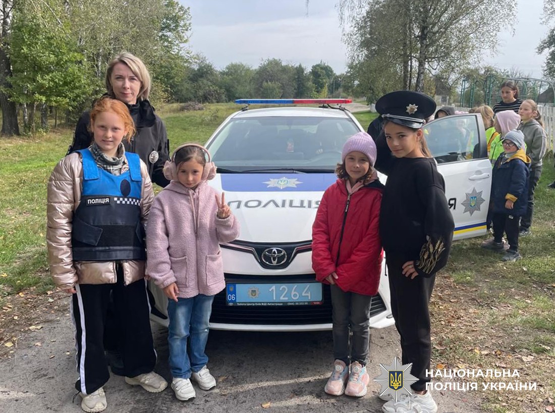 Четверо дівчаток і поліцейська стоять біля службового автомобіля: одна дівчинка у фуражці поліцейського, ще одна у поліцейському жилеті .