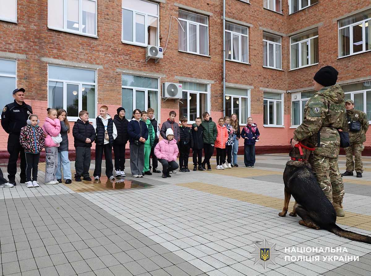На подвір'ї ліцею стоїть група дітей, перед ними поліцейськакінолог зі службовою собакою (вівчаркою)