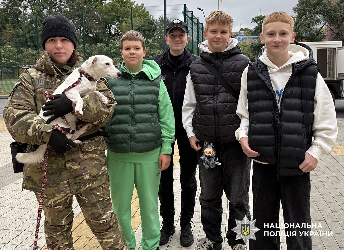 Поліцейська кінолог тримає на руках маленьку білу службову собаку, поруч із нею стоять три ліцеїсти та інспекторка освітньої безпеки, усі позують для фото.