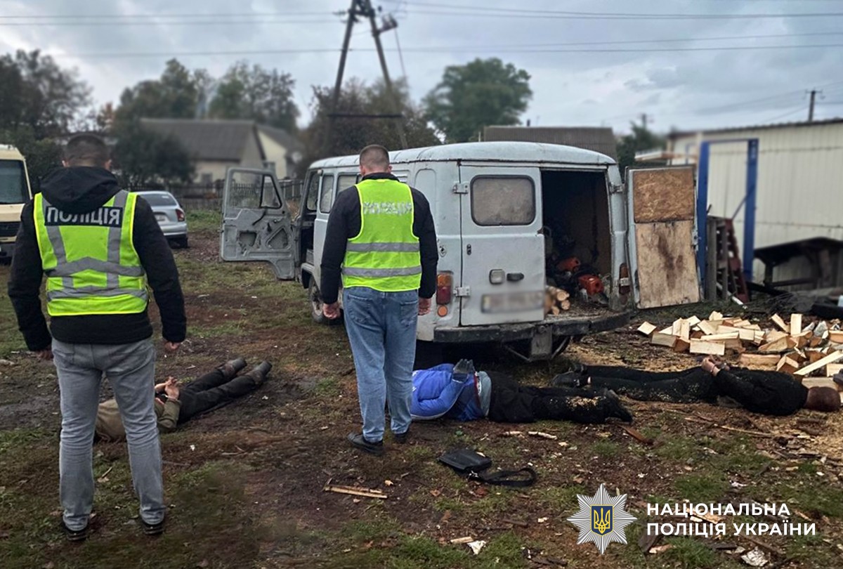 двоє поліцейських затримали трьох фігурантів. правопорушники лежать на землі. навколо будинки, автомобілі, деревина та тирса
